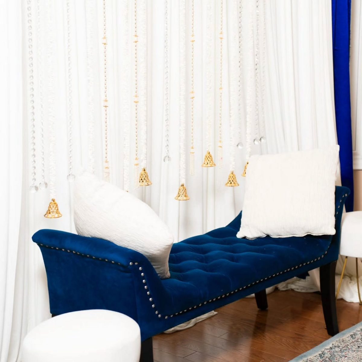 Golden bells up close — close-up of a royal-blue tufted bridal bench with white cushions beneath hanging crystal strands and gold bells; Indian wedding decor rental Toronto