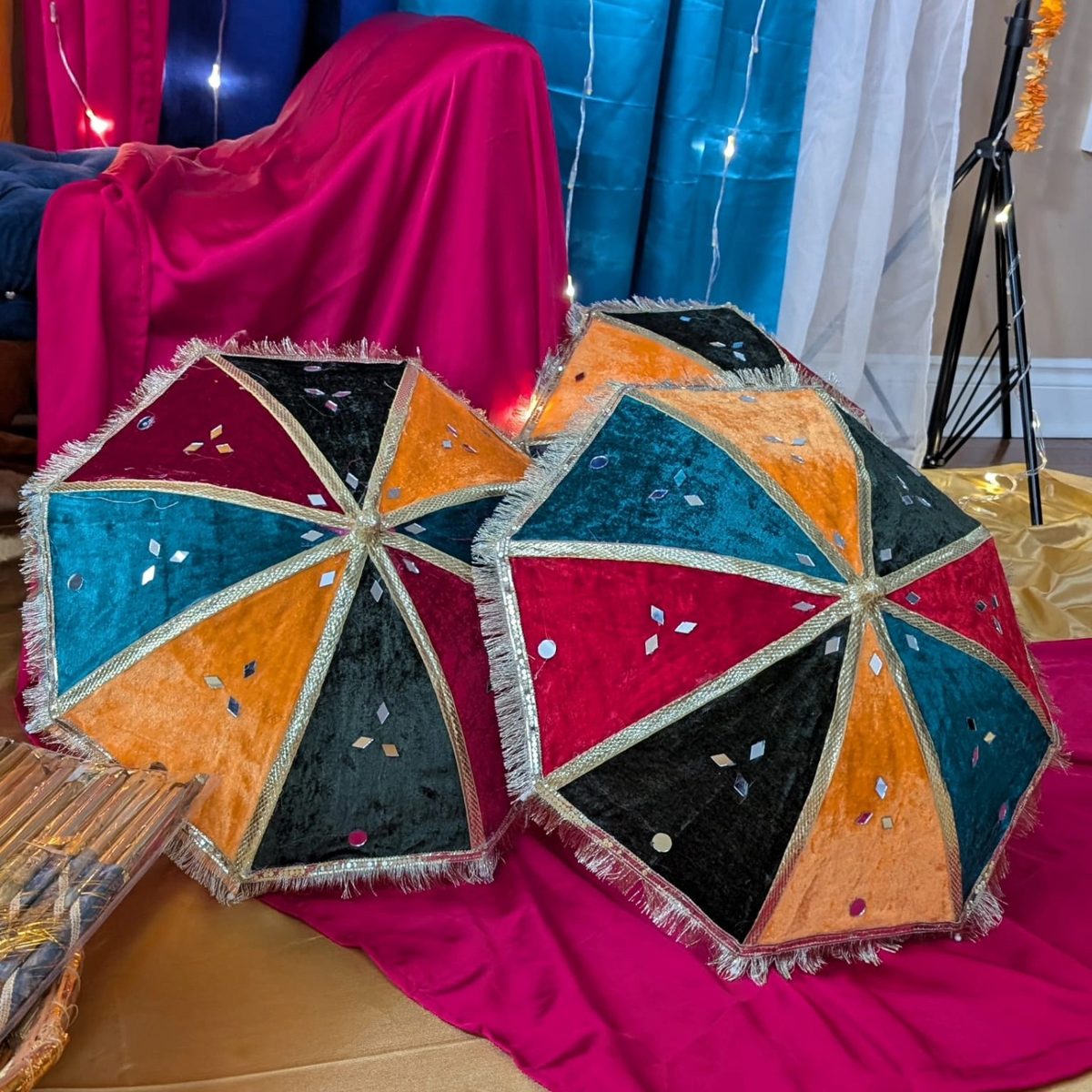 Hand-embroidered mirror-work umbrellas in teal, orange and magenta resting against hot pink and blue drapes — dholki and sangeet decor rental in Toronto