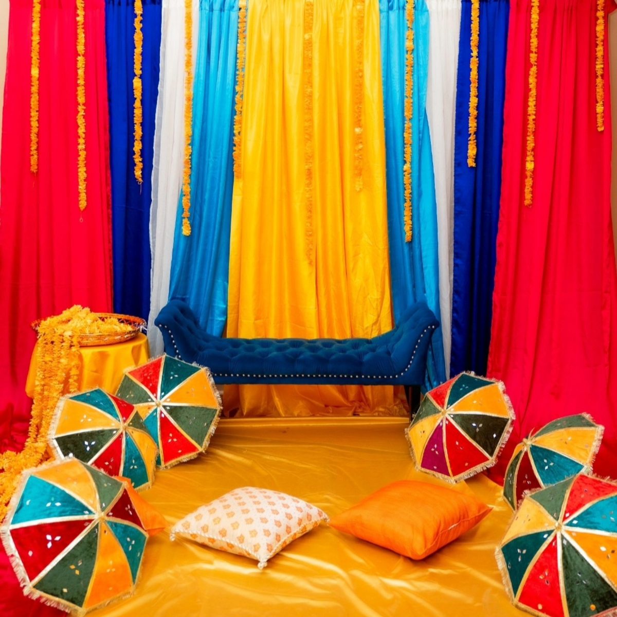 Colour-drenched mehndi stage rental with red, blue and yellow drapes, marigold strands, a navy velvet bridal settee and embroidered umbrellas — Toronto mehndi decoration